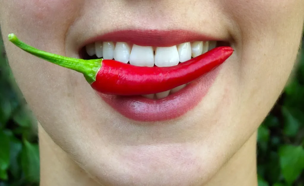 A close-up of a woman’s mouth biting on a piece of red chili pepper