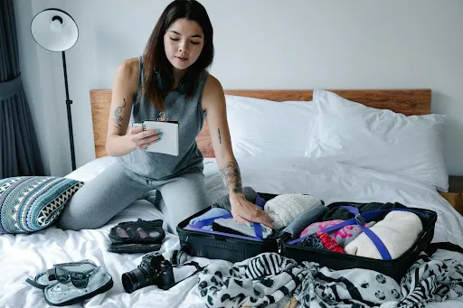 A woman is kneeling on her bed and holding a checklist of her clothes and gadgets