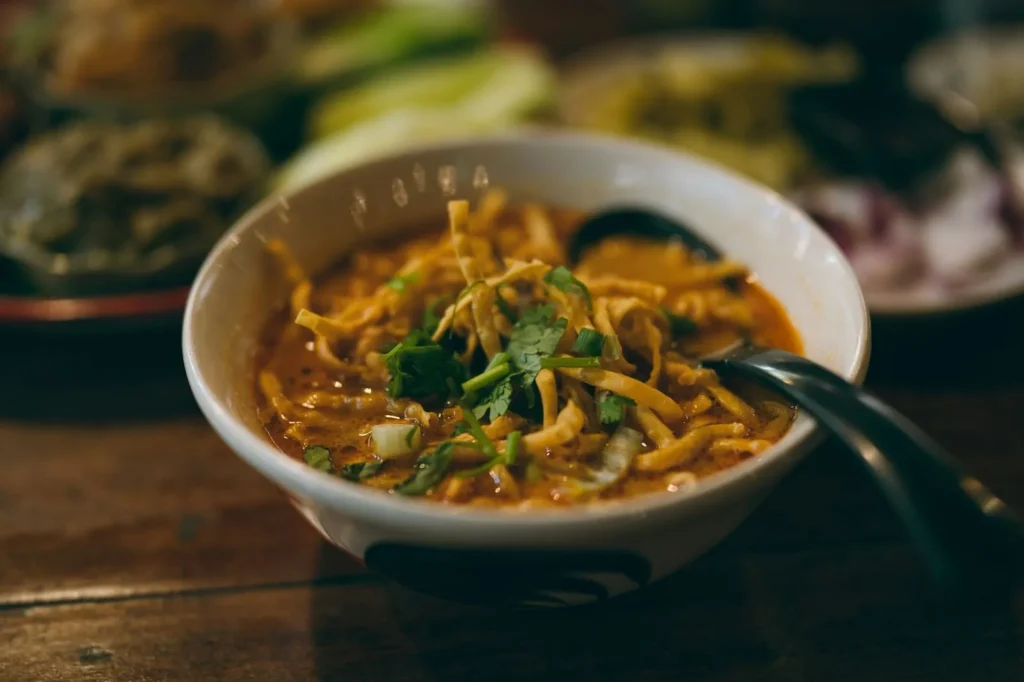 A bowl of Khao Soi on top of a wooden table and served with a black spoon