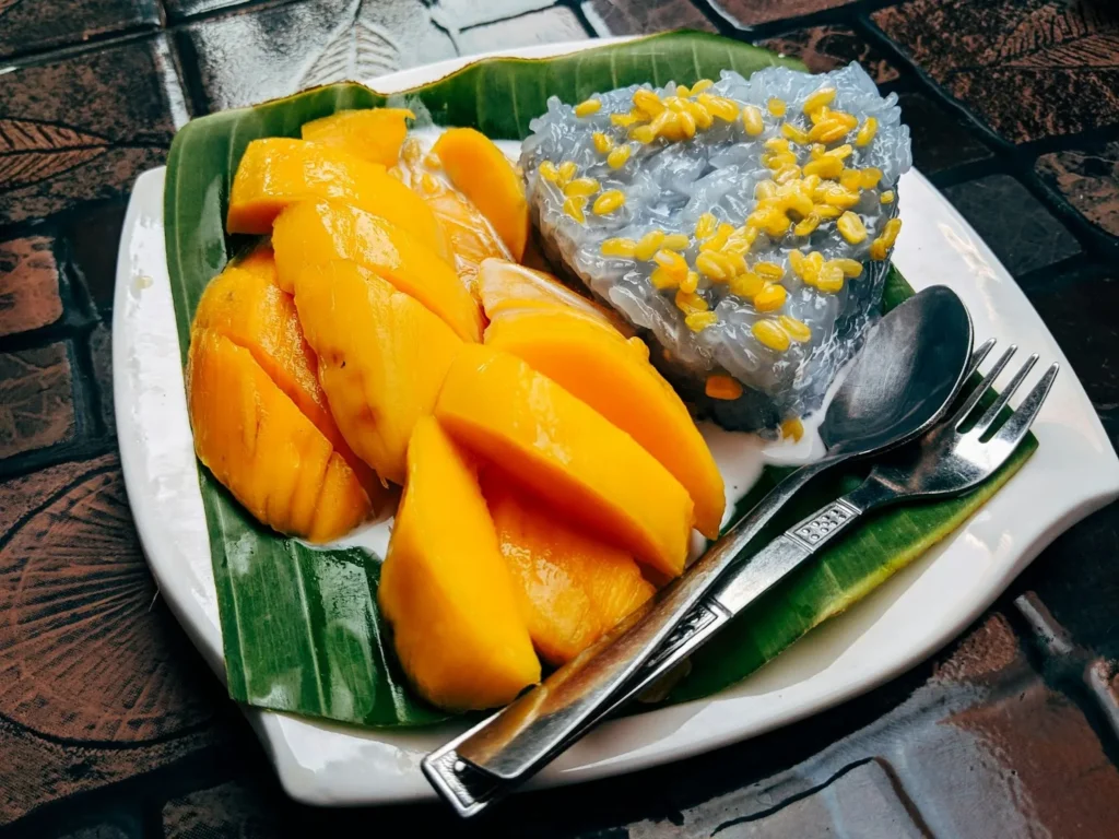 A plate of sliced ripe mangos and sticky rice served on top of a banana leaf  with a spoon and fork on the side