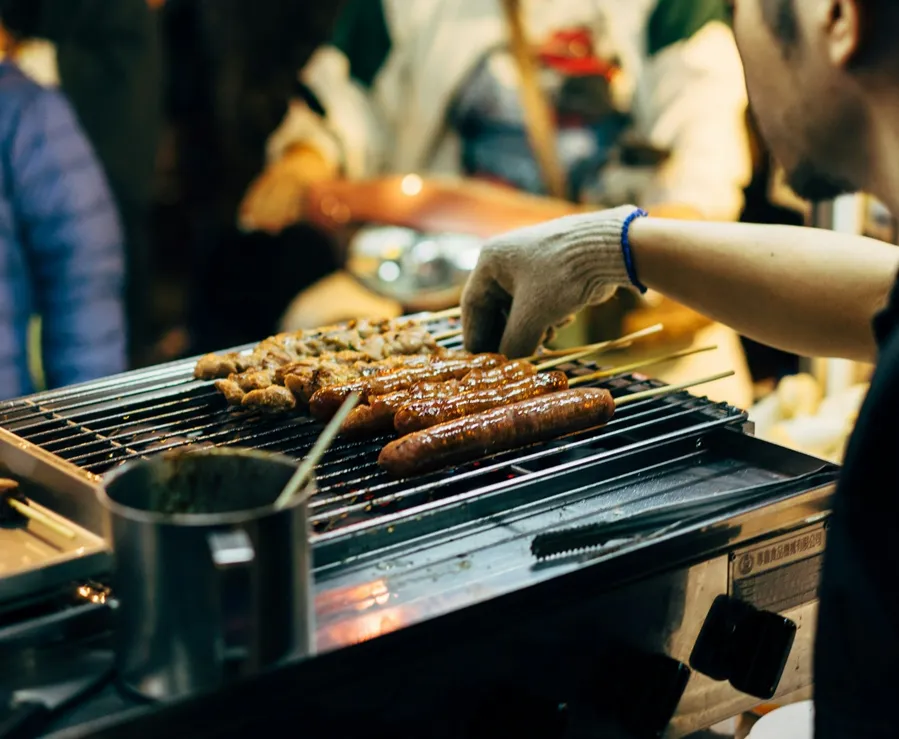 A man wearing gloves is grilling skewered sausages and kebabs on top of a metal griller