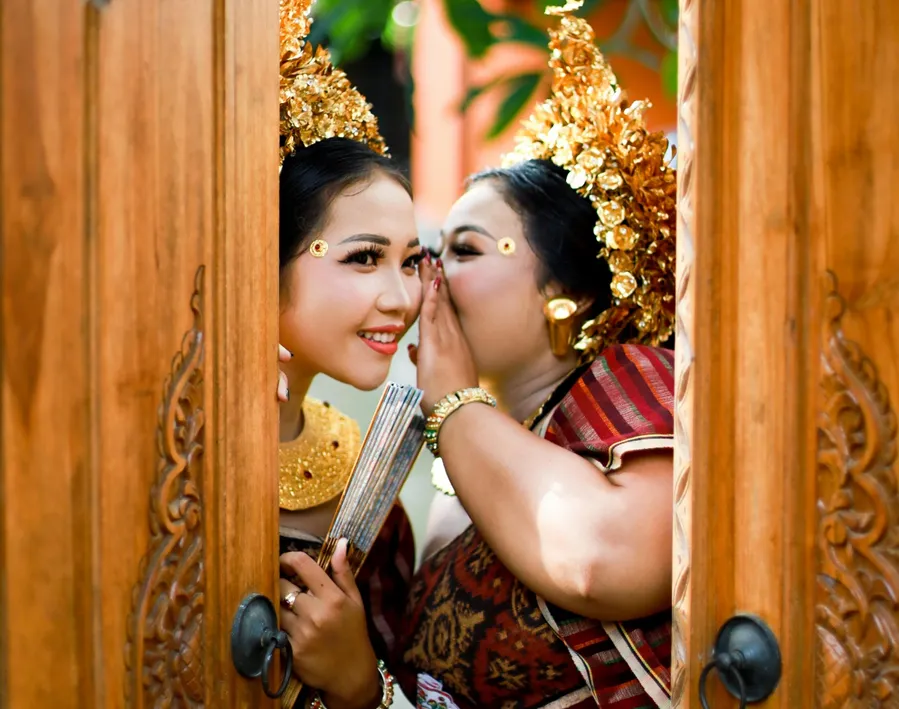 A woman wearing a gold hairdress is whispering to another woman behind a wooden door

